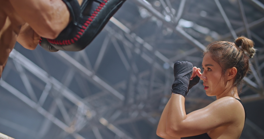 Professional female boxer doing boxing workout punching her instructor trainer in abandoned building dark hall gym. Young asian woman fighter practicing punches training with coach before competition.