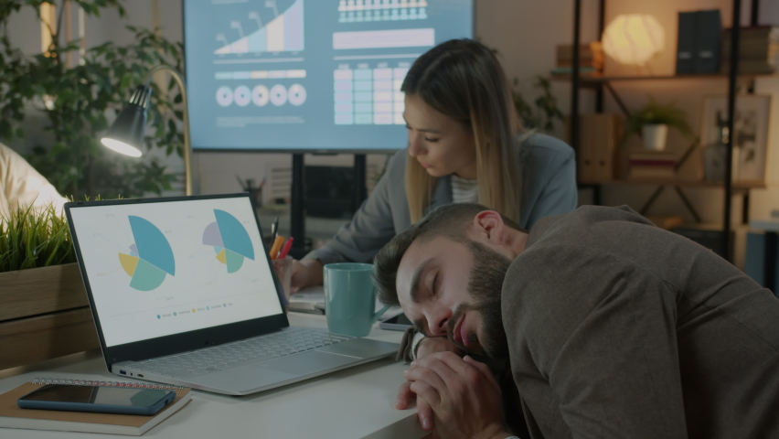 Exhausted employee sleeping on desk in dark office while colleagues working in background. Workplace and tired businesspeople concept. | Shutterstock HD Video #1093054265
