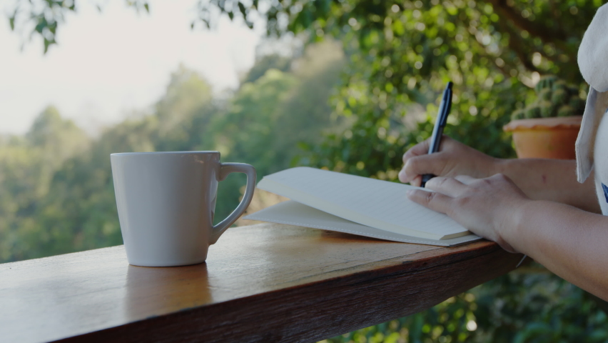 Asian woman writing down in white notebook. Woman sitting on balcony and writing idea on notebooks near fresh nature with cup of coffee in the morning time. | Shutterstock HD Video #1093058683
