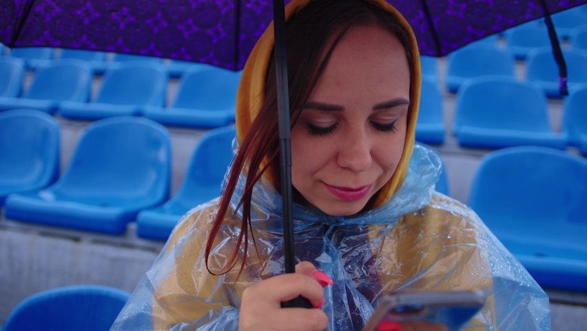 A focused woman in a hoodie and a plastic raincoat using a mobile phone while sitting on the podium with an umbrella in her hand during a match