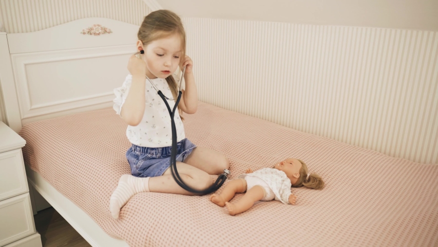 The girl is playing doctor. The child listens to the doll with a stethoscope, sits on the bed.