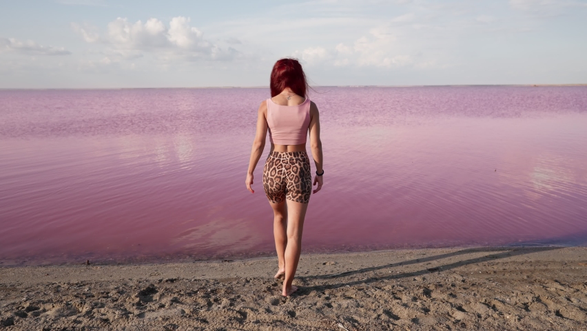 Slim Woman with Red Hair Walks Along Sandy Shore Towards Pink Lake. Water with Health Properties. Travel and Tourist Places Concept. Wellbeing and Active lifestyle. Go Everywhere. Top View Slow