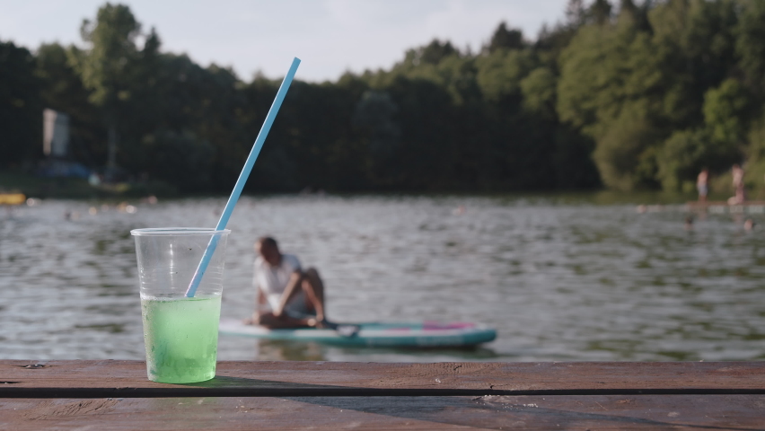 Drink in plastic cup with straw and person on paddleboard in background, summer time or holiday on lake | Shutterstock HD Video #1093071763