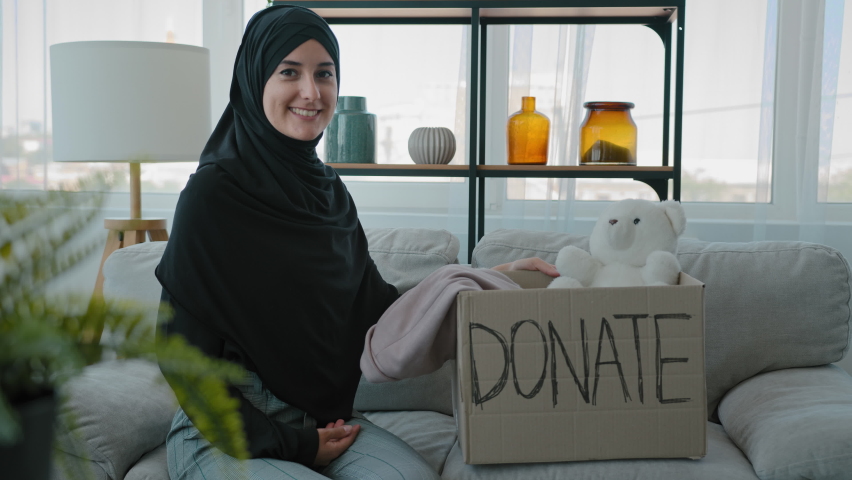 Islamic Muslim woman in black hijab female volunteer help children victims of third world war in Ukraine natural disasters needy poor people sitting on couch packing donations box with clothes and