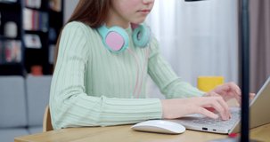Close up on Caucasian teen pretty girl with headphones on neck playing on laptop computer at home. Beautiful schoolgirl tapping and chatting online with smile on face in living room. Indoor.