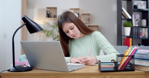 Pretty Caucasian teen girl sitting at laptop, writing exercise in copybook and having online class. School by Internet. Teenage having videochat class by computer at home and doing homework. Learning.