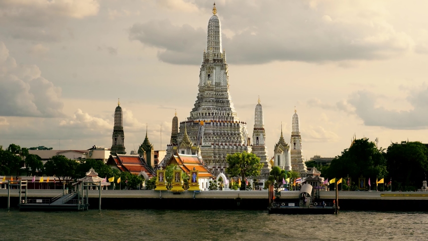 Shot of tourist motor boats docked along the wooden pier over Chao Phraya river with the view of beautiful Wat Arun Temple in background in Bangkok, Thailand at sunset.