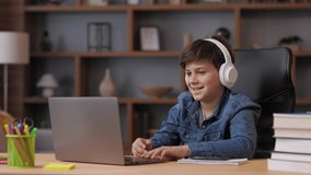 Schoolboy wearing earphones has online lesson, video call with teacher, answering questions, check of knowledge. Cute teenager studying at home using laptop. Distance learning and home education.