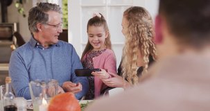 Video of happy caucasian mother, daughter and grandfather looking at smartphone at dinner table. Family, domestic life and togetherness concept digitally generated video.