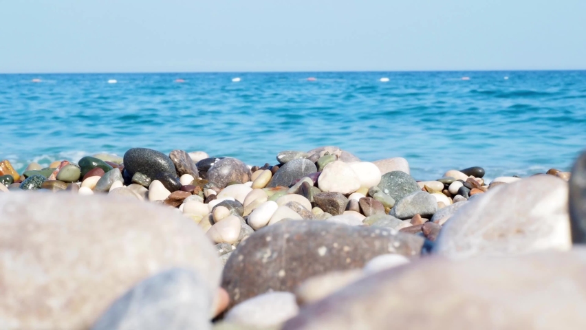 Pebbles on the seashore in the foreground in focus. In the background is the sea with waves. Stones on the beach. Close-up of a sea pebble on the coast | Shutterstock HD Video #1093096823