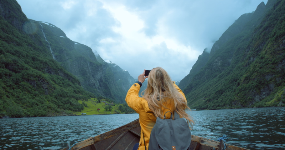 Female travel researcher taking phone photos of green mountain views, sitting alone in a row boat. Back view of woman doing ecology research outside. Traveler takes a picture for online content Royalty-Free Stock Footage #1093098083