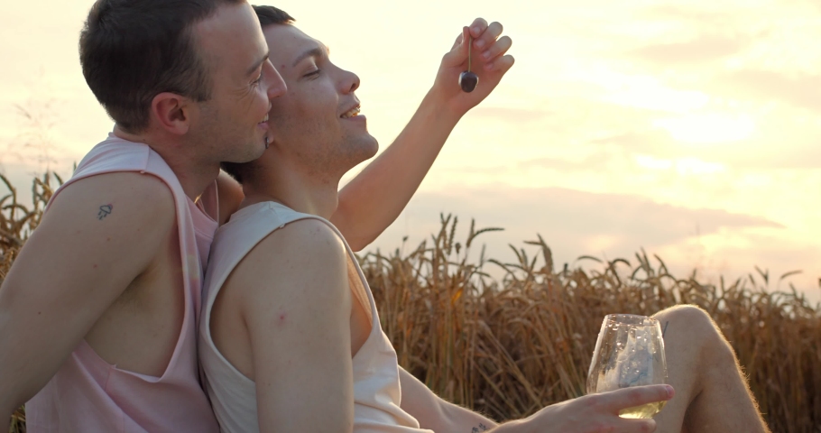Side view of a man feeding his boyfriend with a cherry on the sunset background. Gay couple having fun on a picnic at the wheat field, hugging and resting in nature