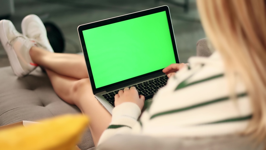 Young blond woman sitting on the sofa at home, holding a green screen laptop on her legs. Over-the-shoulder view of a working woman. | Shutterstock HD Video #1093103851