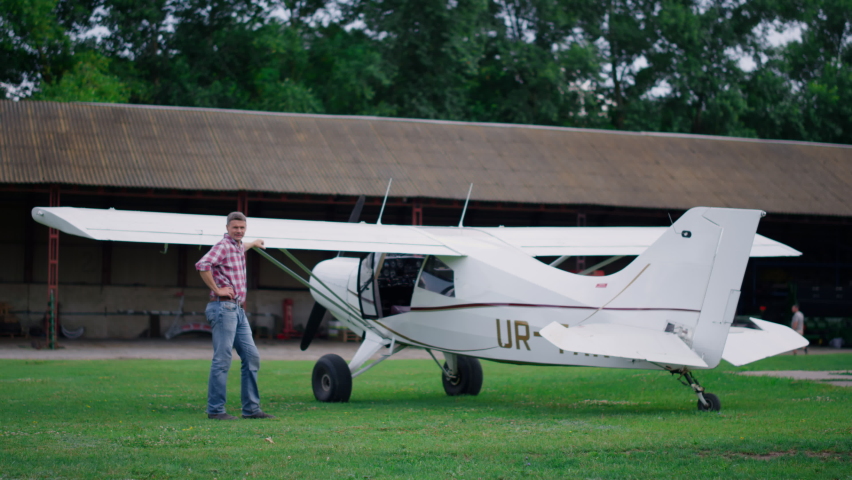 Pilot posing standing near white airplane after successful landing on airfield. Middle age airman leaning on small private plane relaxing after training flight cloudy day. Aviation transport concept