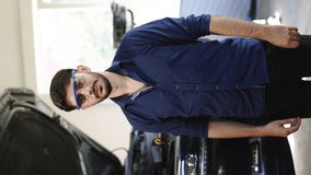 Vertical Format Video of Bearded Handsome Car Mechanic is Posing in a Car Service. He Wears a Jeans Shirt and Safety Glasses. His Arms are Crossed. Specialist Manager Looks at Camera