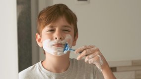 Boy in front of mirror in bathroom removes shaving foam from his face with razor. Video portrait of child imitating dads shaving.
