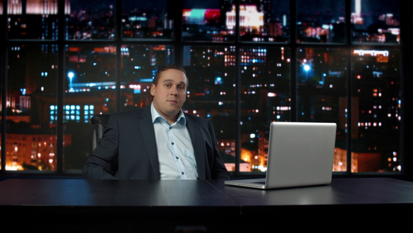 A man in a business suit, a TV news and show, sits at a desk in a studio with a laptop and background night city.