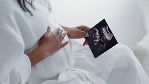 Maternity love. Close up shot of unrecognizable african american pregnant woman admiring sonography picture of her unborn baby and caressing belly, sitting at home, slow motion - Βίντεο στοκ