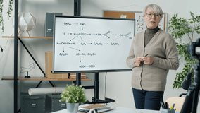 Experienced Chemistry teacher recording tutorial for science blog with video camera. Mature woman using digital screen to show formula and speaking.