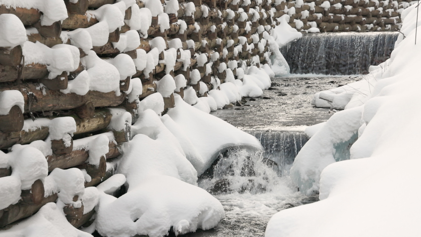 Front view of small cascade waterfall falling down in winter mountine forest. Slow motion of water stream flowing down along snowy shores with pattern wooden dam on background. Concept of landscape.