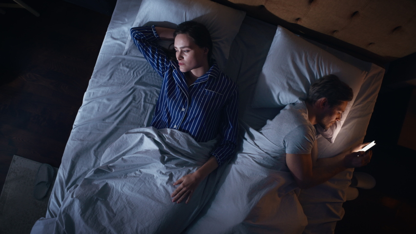 Top View Bedroom Apartment: Man Uses Smartphone in Bed at Night When His Female Partner Trying to Fall Asleep. Couple After Fight, Argument. Addictive World of Social Media, Doom Scrolling, Fake News. Royalty-Free Stock Footage #1093149705