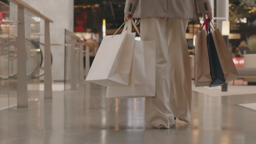 Low section rear view tracking slowmo of unrecognizable young woman in smart casualwear carrying many paper shopping bags walking confidently towards food court in modern shopping center