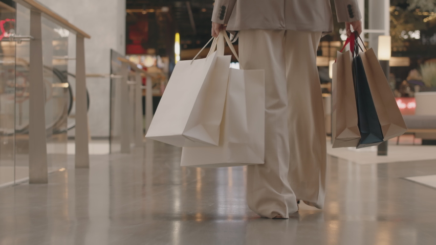 Low section rear view tracking slowmo of unrecognizable young woman in smart casualwear carrying many paper shopping bags walking confidently towards food court in modern shopping center Royalty-Free Stock Footage #1093151715