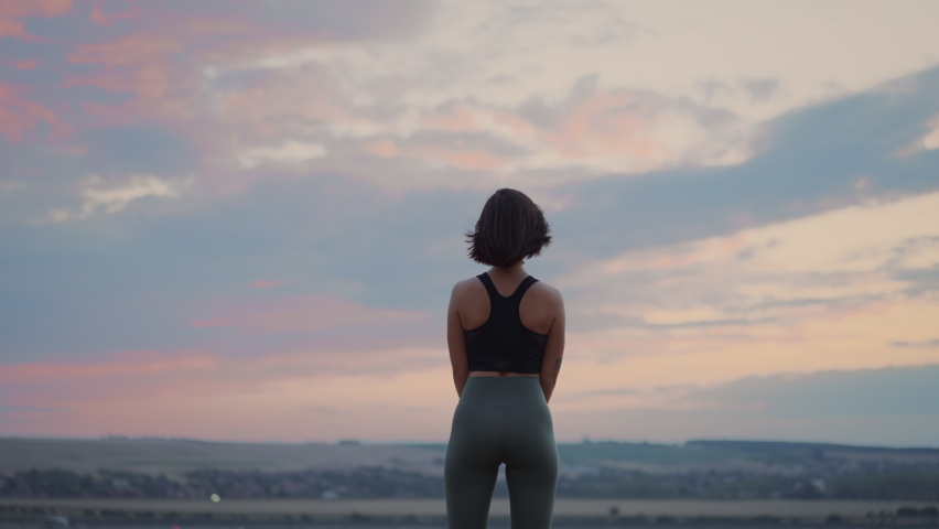 Back view of the beautiful woman wearing sports clothing relaxing and deeply breathing on the hill on the grass