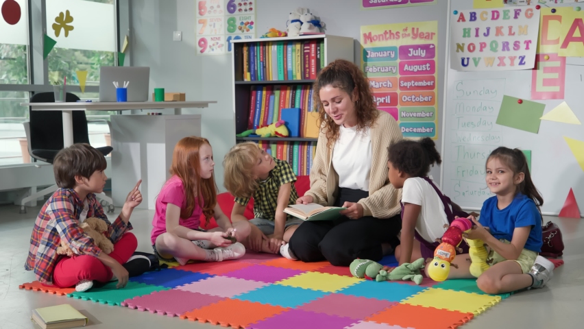 Female teacher reading story to group of elementary pupils in school classroom. Young teacher and preschool kids sit on classroom floor and read book together in kindergarten | Shutterstock HD Video #1093177729