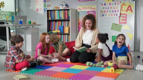 Стоковое видео: Female teacher reading story to group of elementary pupils in school classroom. Young teacher and preschool kids sit on classroom floor and read book together in kindergarten