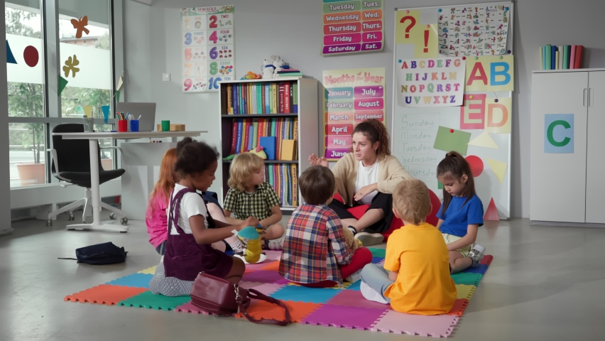 Small nursery school children with teacher sitting on floor having lesson. Young woman teach preschool kids in classroom sitting on floor together | Shutterstock HD Video #1093177747