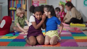 Multiracial little girls tell secrets sitting on floor in kindergarten. Adorable preschool African-American and caucasian girls sitting together in playroom share secrets - Powered by Shutterstock - Get 15% off with code: PIKWIZARD15
