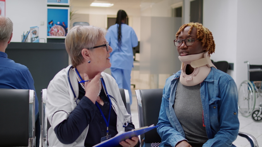 Old medic consulting african american woman with cervical collar, sitting in hospital reception waiting area. Physician doing checkup examination with injured patient wearing neck foam. | Shutterstock HD Video #1093184203