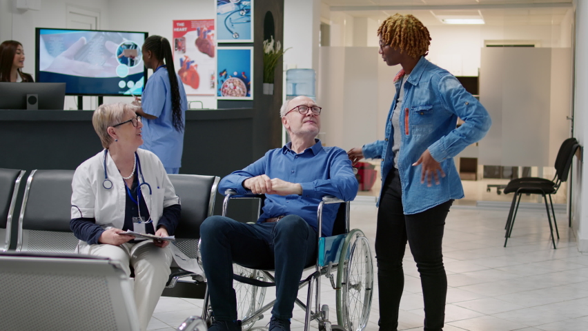 Man with physical impairment attending medical checkup with doctor and caretaker in waiting area. Patient wheelchair user talking to physician at clinic, having social assistance carer. | Shutterstock HD Video #1093184207