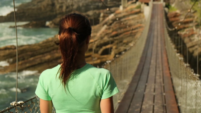 Sporty woman running on mountain road in beautiful nature. A brunette runner woman runs in the park jogging. sports girl back view runs in the morning on a suspension bridge near the ocean, america
