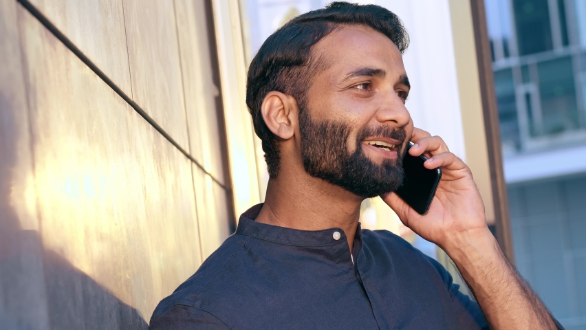 Smiling young indian business man holding smartphone talking on cell phone making call standing on urban street in modern city on sunset speaking by cellphone, laughing, having mobile conversation. Royalty-Free Stock Footage #1093185905