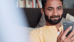 Happy indian man user sitting at home relaxing on sofa wearing earbud using smartphone looking at cell phone watching social media videos, listening music podcast, having virtual mobile chat call. - Powered by Shutterstock - Get 15% off with code: PIKWIZARD15