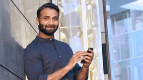 miling indian professional business man wearing earbud holding cellphone using mobile phone watching video, having online chat call, using apps standing in urban city on modern street outdoors.