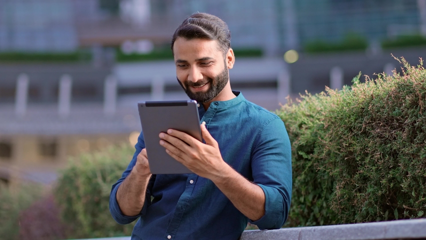 Smiling happy young indian business man professional, eastern businessman executive standing outdoors in urban city park using digital tablet pad online technology browsing surfing internet outside. Royalty-Free Stock Footage #1093185925