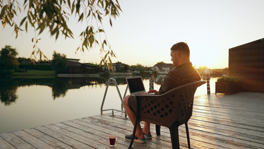 Young man relaxing on a wooden deck at sunset in a comfy chair typing on his laptop computer