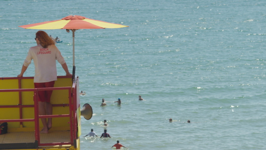 Lone lifeguard looks out to sea as people play in the waters, in slow motion - space for text Royalty-Free Stock Footage #1093186953
