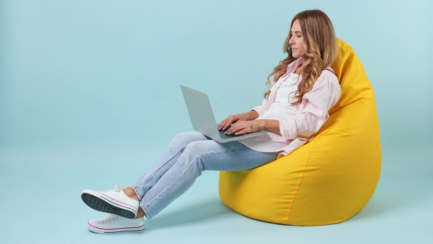 Side view full body young happy it woman wear pink shirt sit in bag chair hold in hand type use work on laptop pc computer isolated on plain pastel light blue cyan background. People lifestyle concept