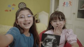 Little girls holding an image of their unborn sibling from ultrasound. Girls take selfie with ultrasound photo of their pregnant mother, describe surprise on video call.