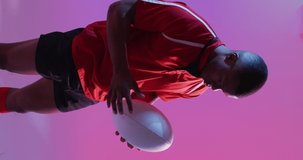 Vertical video of african american male rugby player with rugby ball over pink lighting. Sport, movement, training and active lifestyle concept.