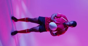 Vertical video of portrait of african american male rugby player with rugby ball over pink lighting. Sport, movement, training and active lifestyle concept.