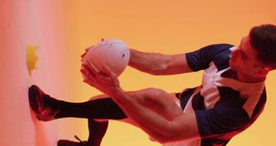 Vertical video of caucasian male rugby player crouching with rugby ball over pink lighting. Sport, movement, training and active lifestyle concept.