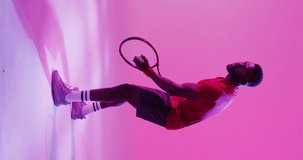 Vertical video of african american male tennis player hitting ball on neon pink background. Professional sport, training and vintage style concept.