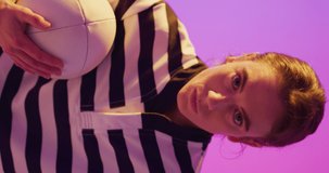 Vertical video of portrait of caucasian female rugby player with rugby ball over neon pink lighting. Sport, movement, training and active lifestyle concept.