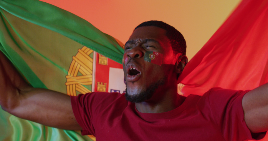Happy african american male football supporter with flag of portugal over pink lighting. Sport, movement, training and active lifestyle concept.