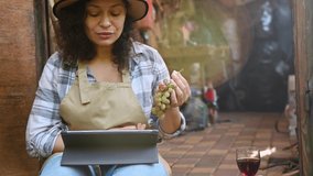 Close-up of a beautiful woman in a beige apron and straw hat, sitting on the doorstep of her workshop, presenting an assortment of grapes grown in her own vineyards while having a video call on tablet